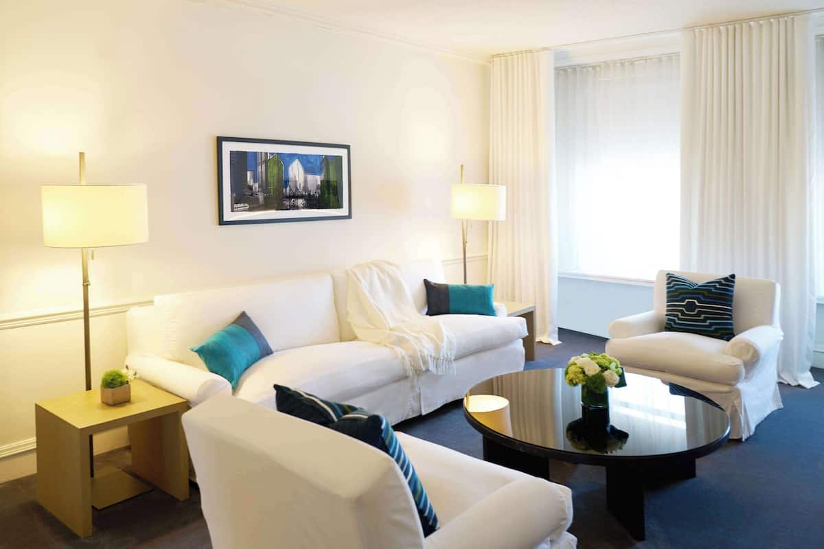 A modern hotel room with a white sofa, a round coffee table, and a white armchair. There is a framed picture on the wall and a floor lamp on a side table.
