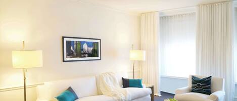 A modern hotel room with a white sofa, a round coffee table, and a white armchair. There is a framed picture on the wall and a floor lamp on a side table.
