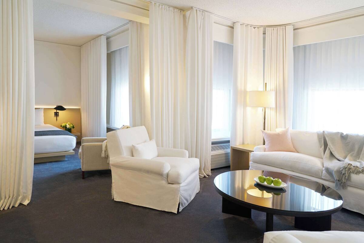 A hotel room with a bed, two white armchairs, a round coffee table with a fruit plate, a sofa, and a lamp.
