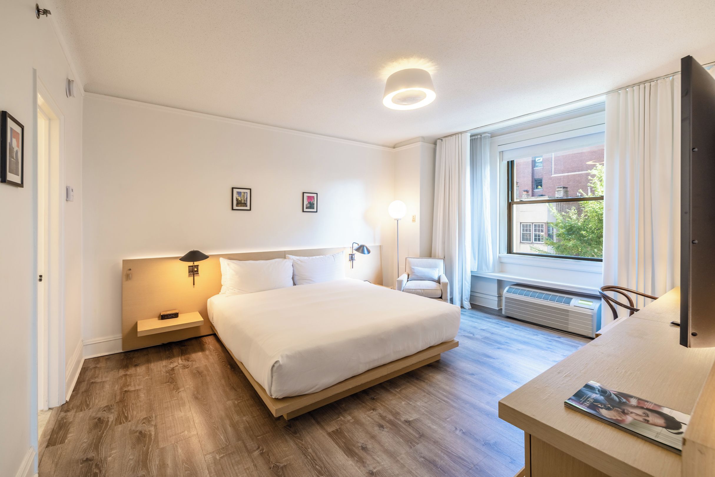 A hotel room with a large bed, bedside tables, a desk with a chair, a window with curtains, and a radiator.