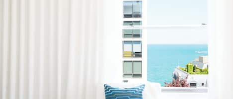 A white armchair with a blue patterned pillow, a window with a view of the sea and buildings, and white curtains.