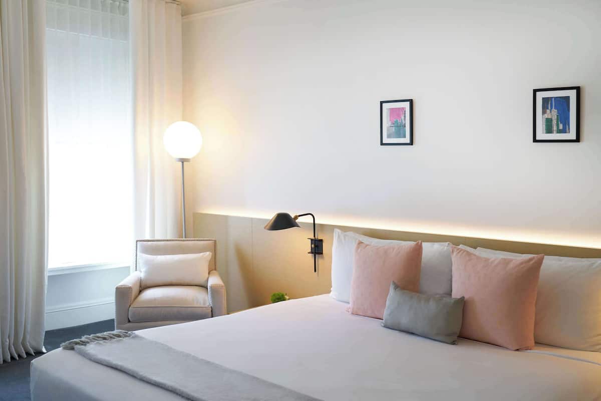 A hotel room with a bed, a chair, a lamp, and two framed pictures on the wall.