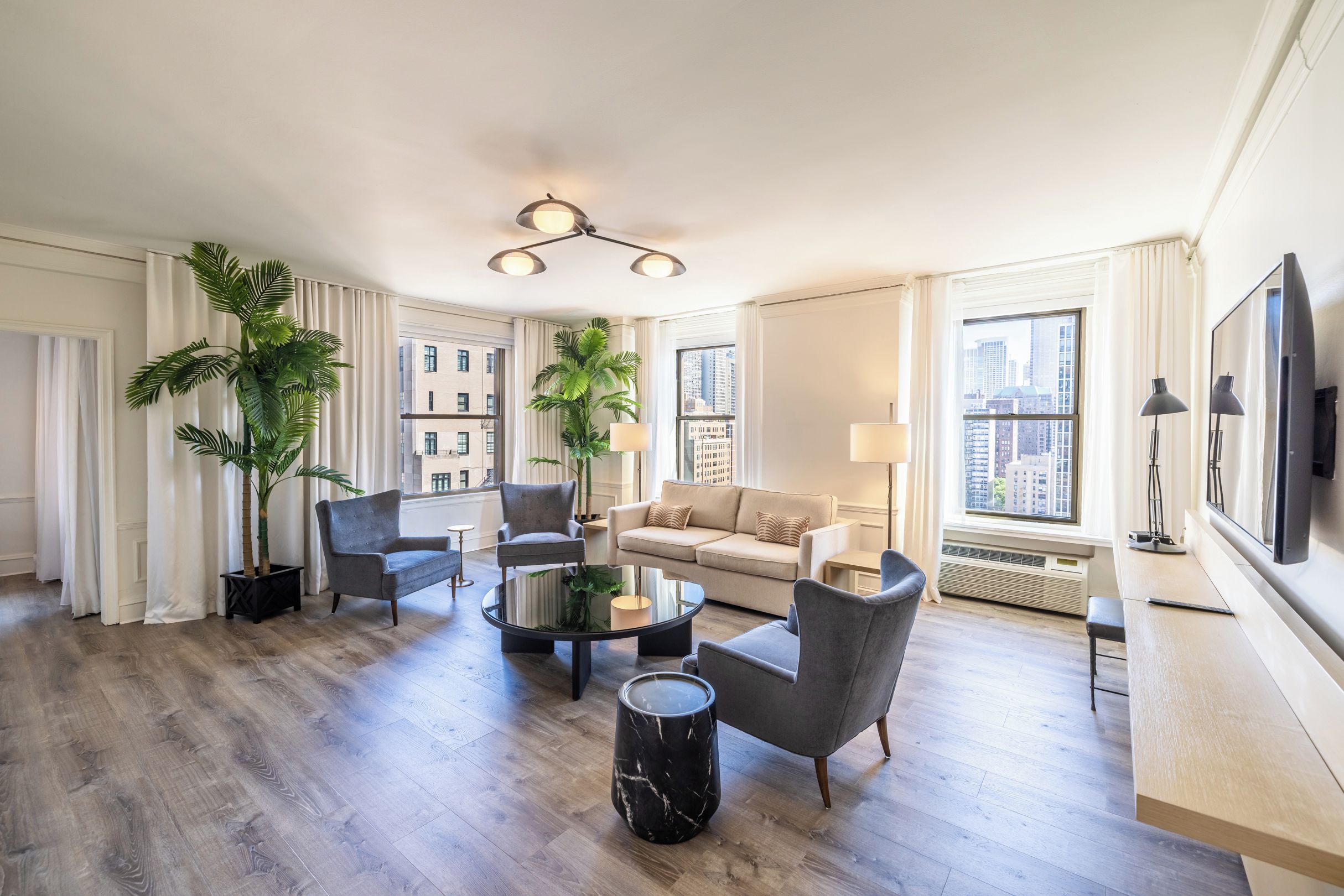 A spacious living room with a round coffee table, two armchairs, a sofa, and a large window offering a city view.