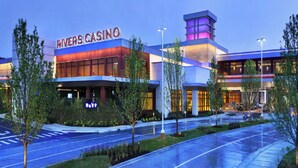 En modern kasinobyggnad, Rivers Casino, med en framträdande skylt, omgiven av välskötta grönområden och en våt väg, vilket troligen tyder på att det nyligen har regnat.