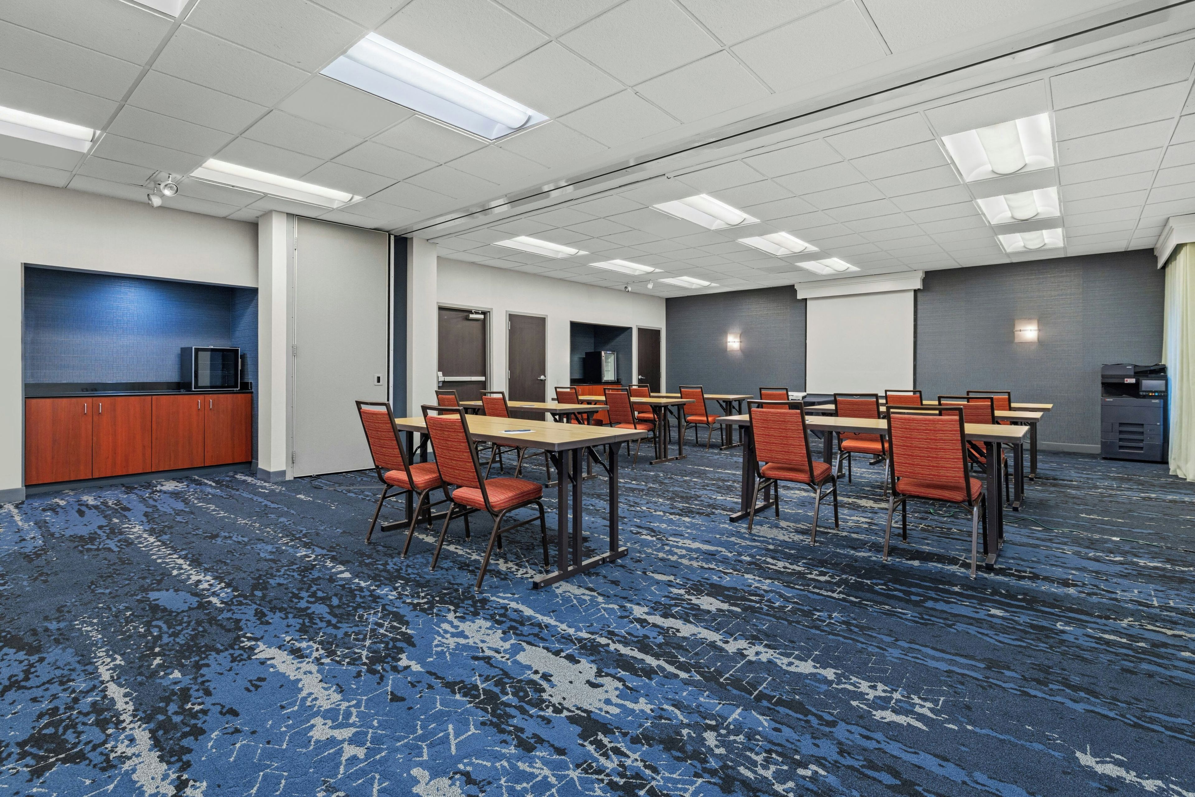 A conference room with multiple tables and chairs, a television, and a kitchenette area.