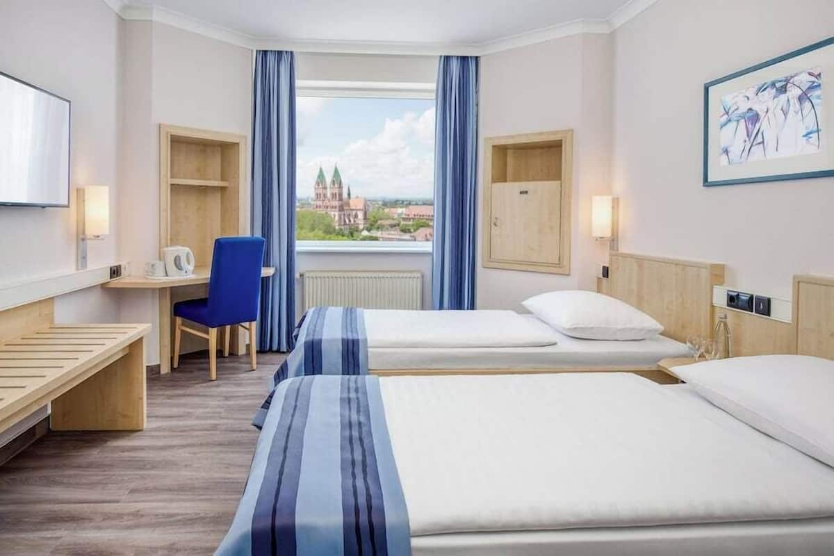 A hotel room with two beds, a desk, a chair, a TV, and a window with a view of a building.