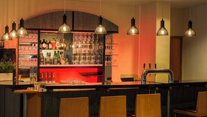 Zona de bar con sillas de madera, una barra con botellas y vasos, e iluminación colgante.
