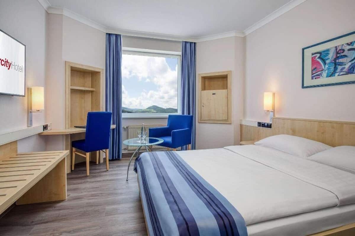 A hotel room with a bed, a desk, a chair, a small table, a window with curtains, and a painting on the wall.