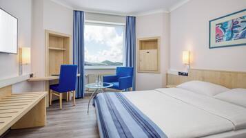 Habitación de hotel con cama, escritorio, dos sillas, una mesita, ventana con cortinas, armario de madera y un cuadro en la pared.
