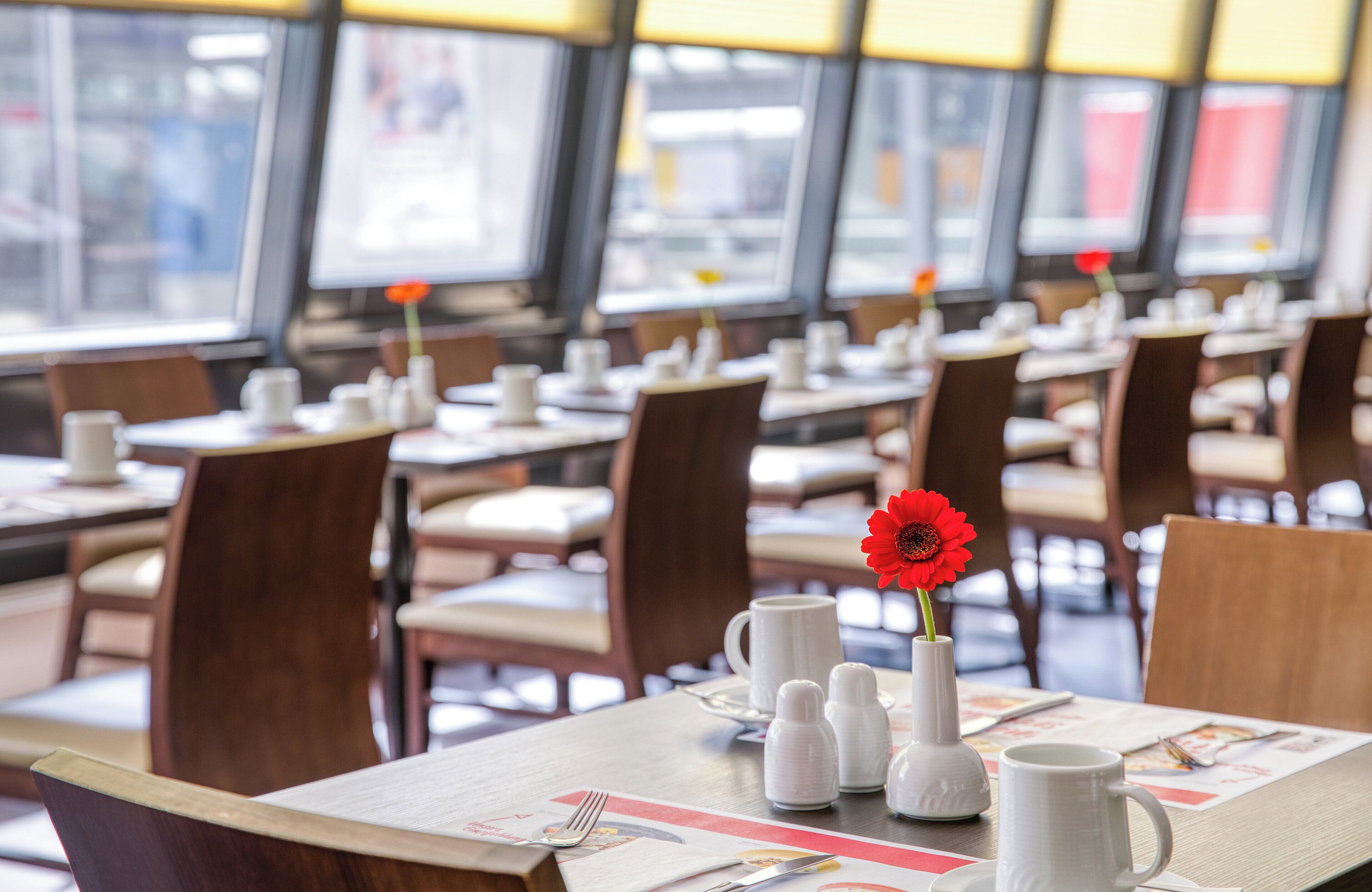 A dining area with tables set for meals, chairs, and a vase with a red flower.