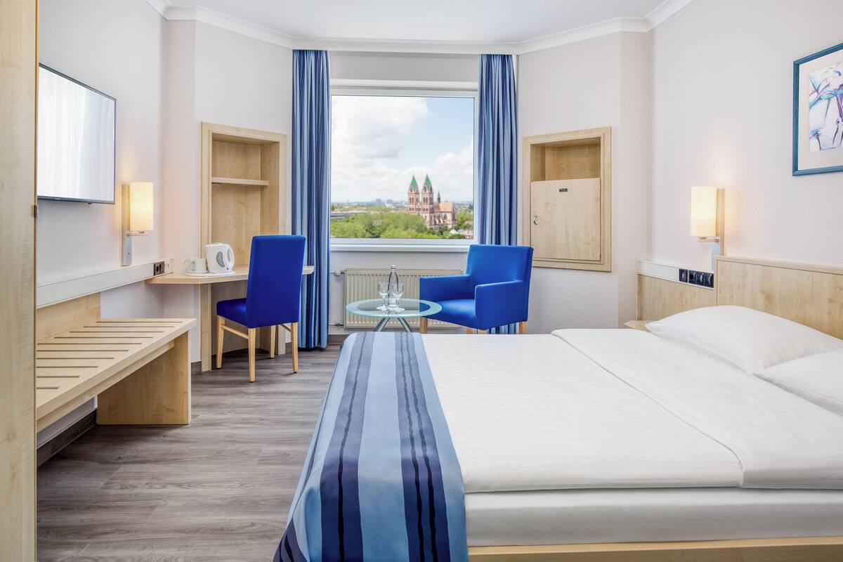 A hotel room with a bed, two blue chairs, a small table with a glass, a wooden desk, a TV, and a window with a view of a building.