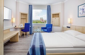 Una camera d'albergo con un letto, due poltrone blu, un tavolino con un bicchiere, una scrivania in legno, una televisione e una finestra con vista su un edificio.