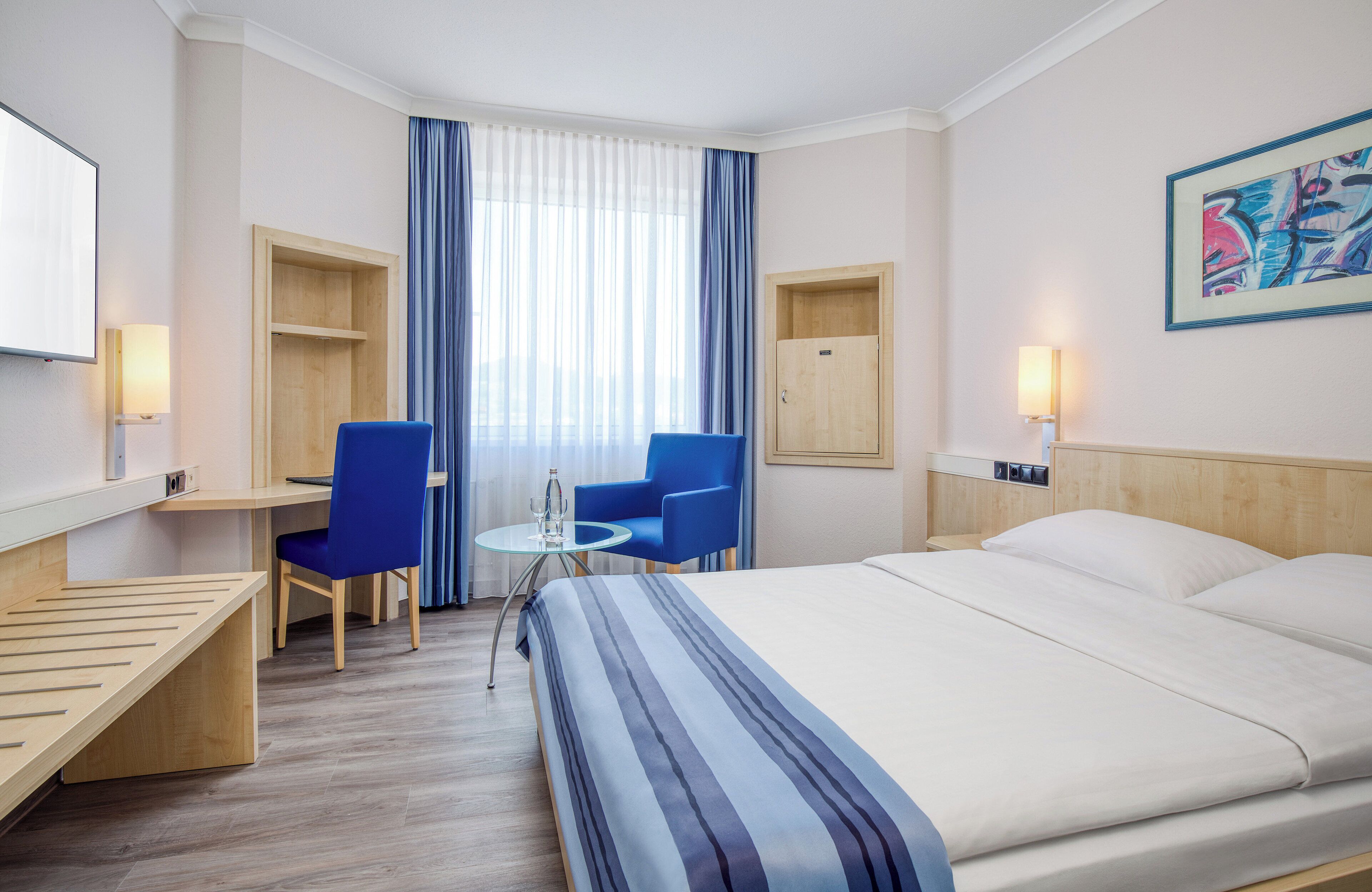 A hotel room with a bed, a desk, two chairs, a small table, a TV, and a window with curtains.