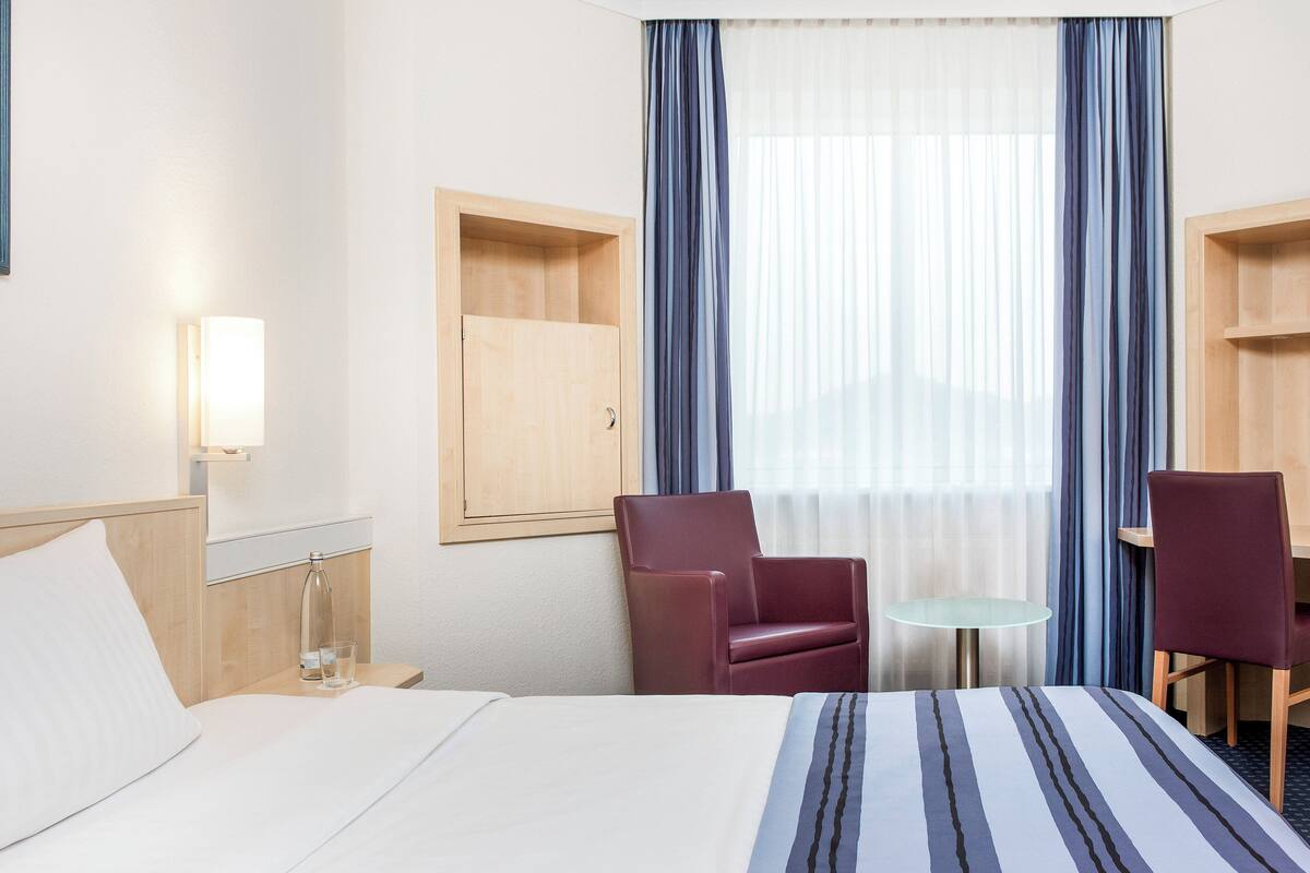 A hotel room with a bed, a desk, two chairs, a small table, a nightstand, a lamp, and a window with curtains.
