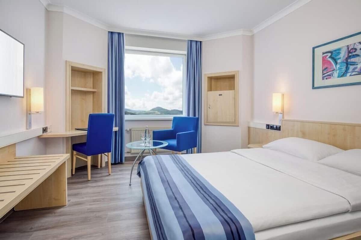 A hotel room with a bed, a desk, two chairs, a small table, a window with curtains, and a painting on the wall.
