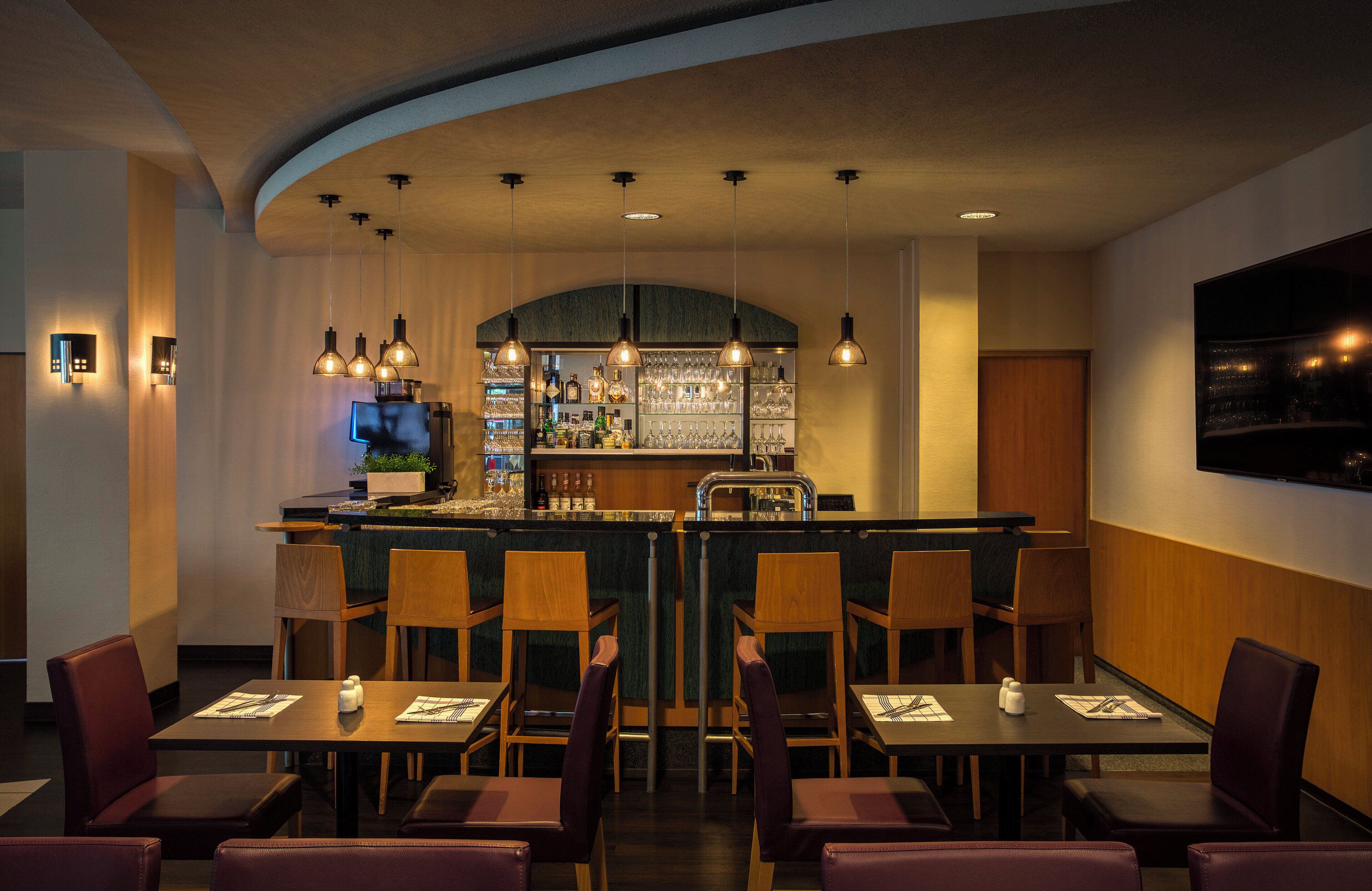 A bar area with a curved ceiling, pendant lighting, and a bar counter with shelves of bottles and glasses. There are several wooden tables and chairs arranged for dining.