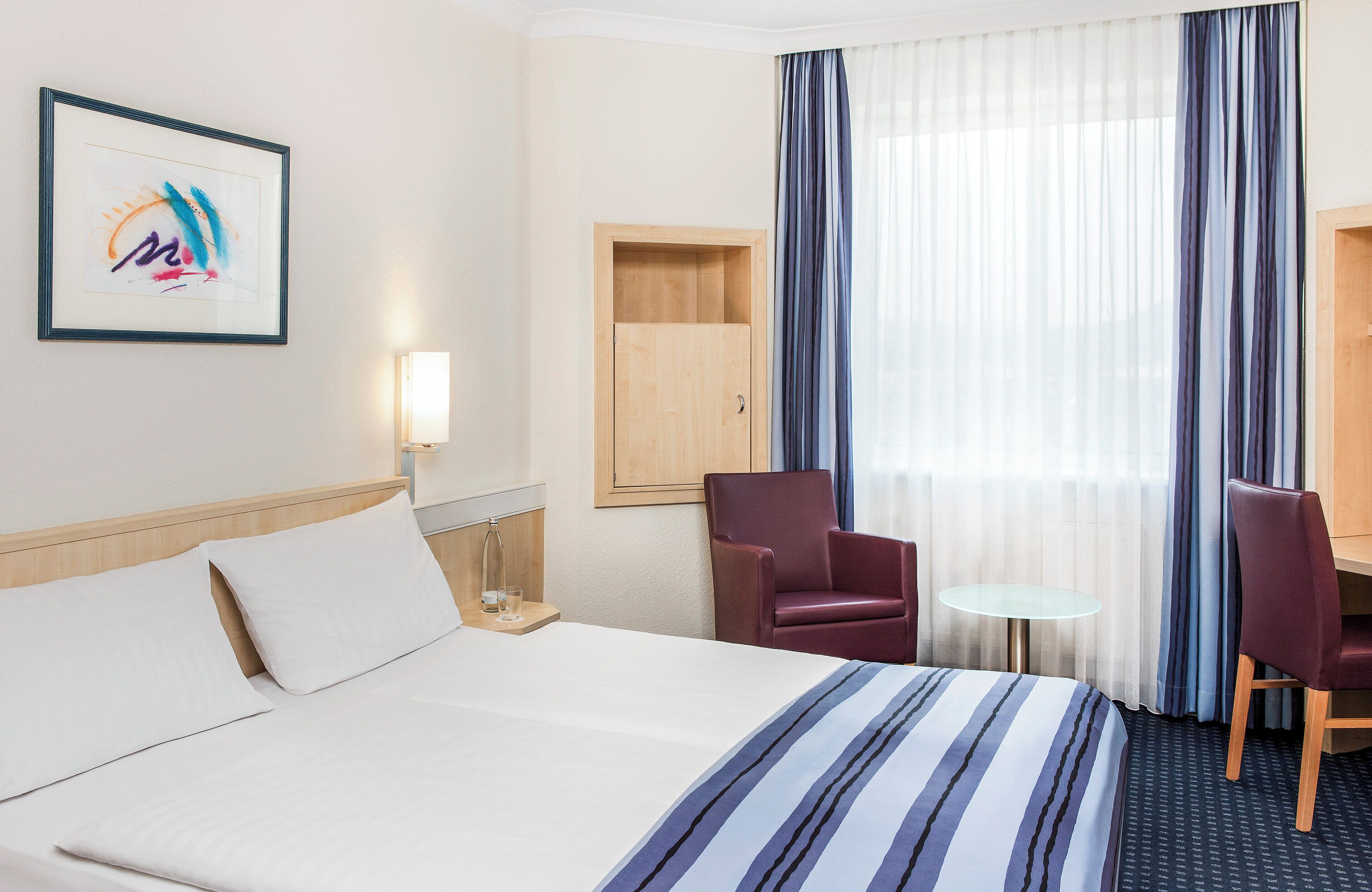 A hotel room with a bed, two chairs, a small table, a lamp, and a window with curtains.