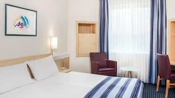 Habitación de hotel con cama, dos sillas, una mesita, lámpara y ventana con cortinas.