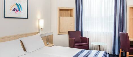 A hotel room with a bed, two chairs, a small table, a lamp, and a window with curtains.