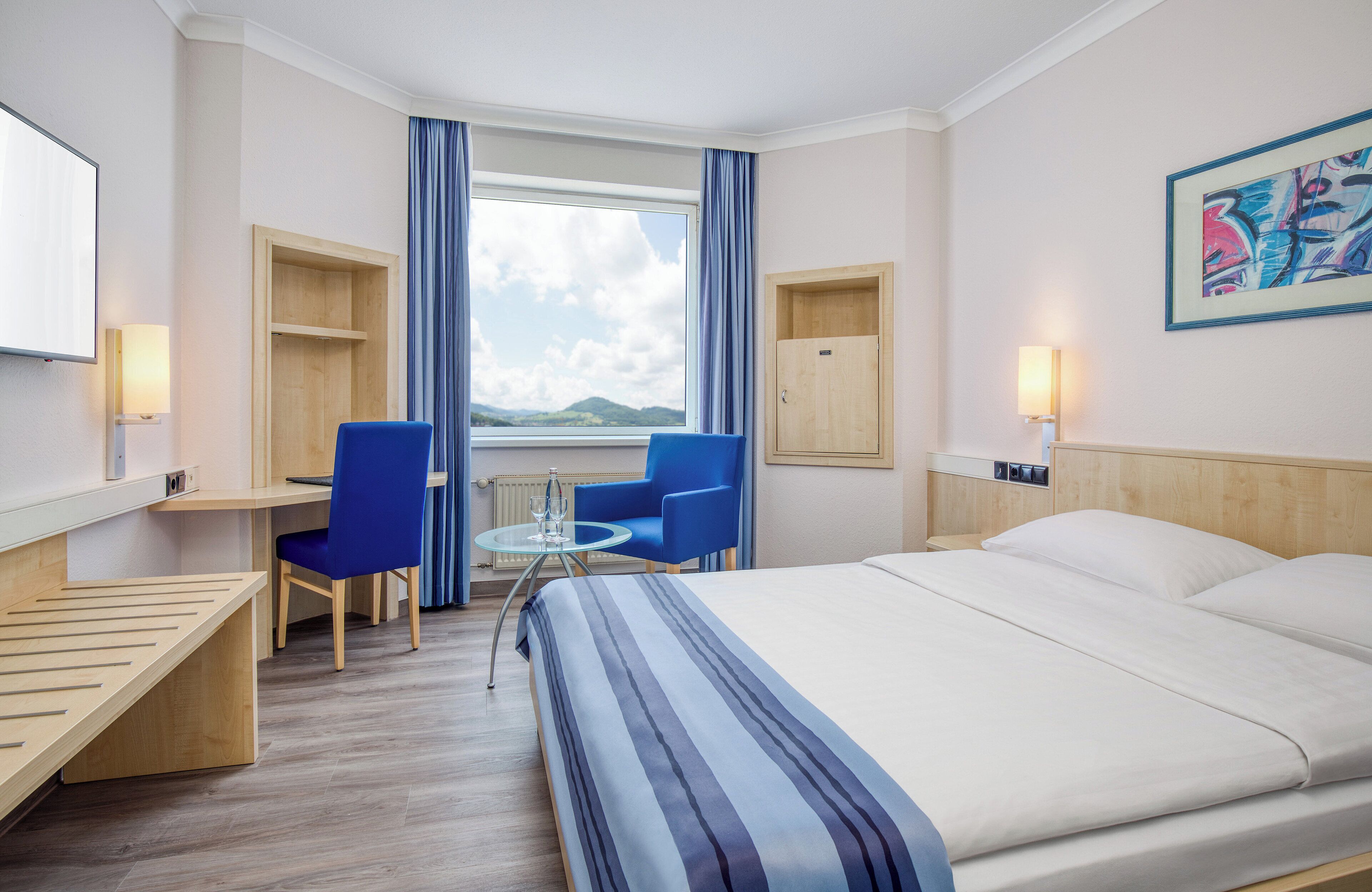 A hotel room with a bed, a desk, a chair, a small table, and a window with curtains.