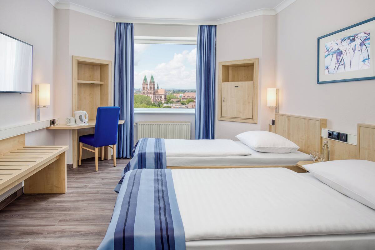 A hotel room with two beds, a desk with a chair, a TV, and a window with a view of a church.