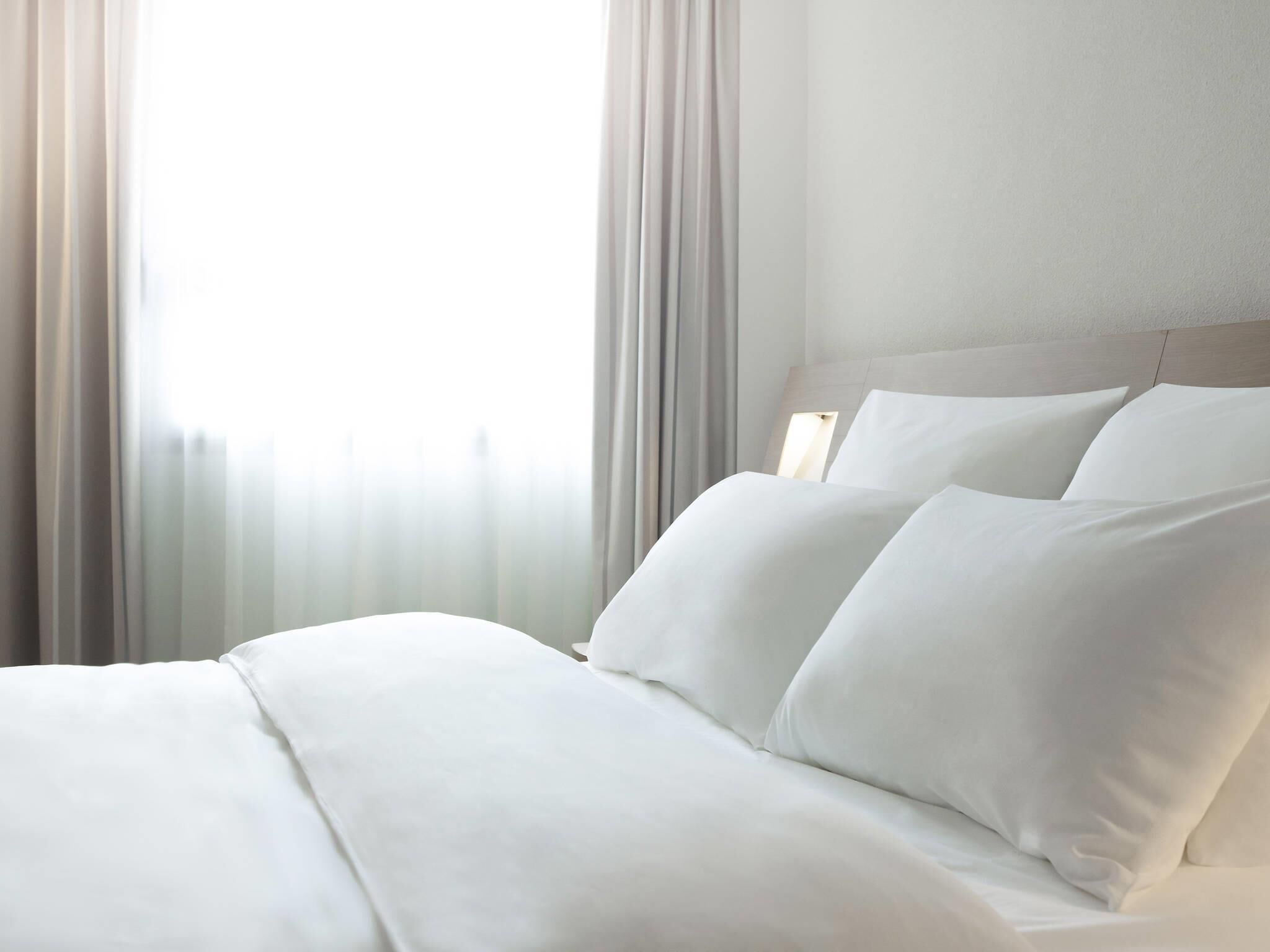 Ein Hotelzimmer mit einem ordentlich bezogenen Bett, einem Kopfteil und durchsichtigen Vorhängen.
