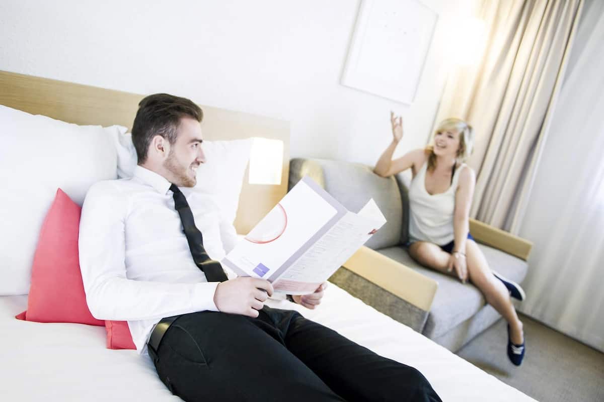 Een hotelkamer met een bed, een bank, een rode kussen, een witte muur en een raam met gordijnen.