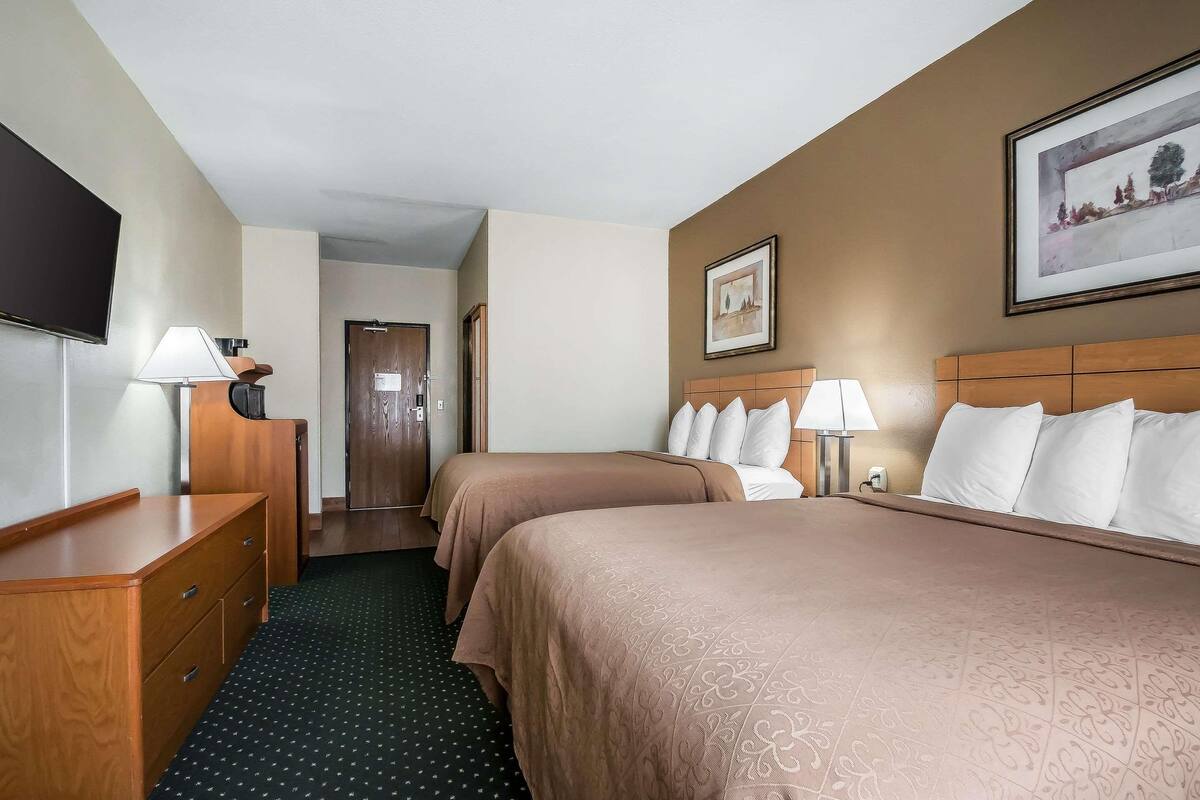 A hotel room with two beds, a desk, a TV, a door, and a framed picture on the wall.