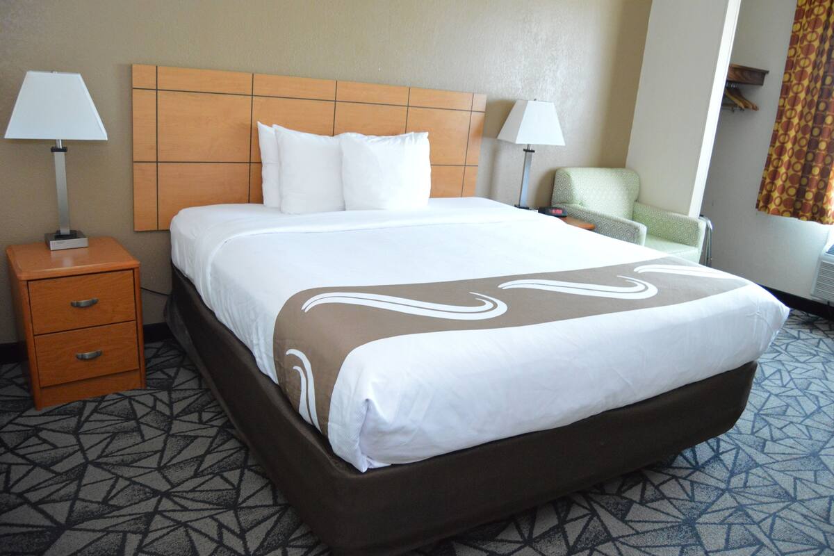 A hotel room with a large bed, two bedside tables with lamps, a chair, and a patterned carpet.
