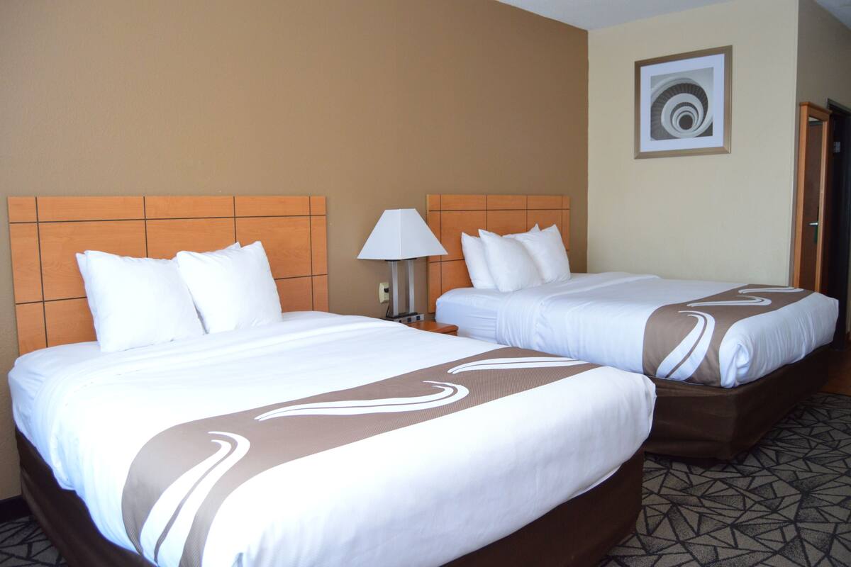 A hotel room with two beds, each with white bedding and brown accents, a nightstand with a lamp, and a framed artwork on the wall.