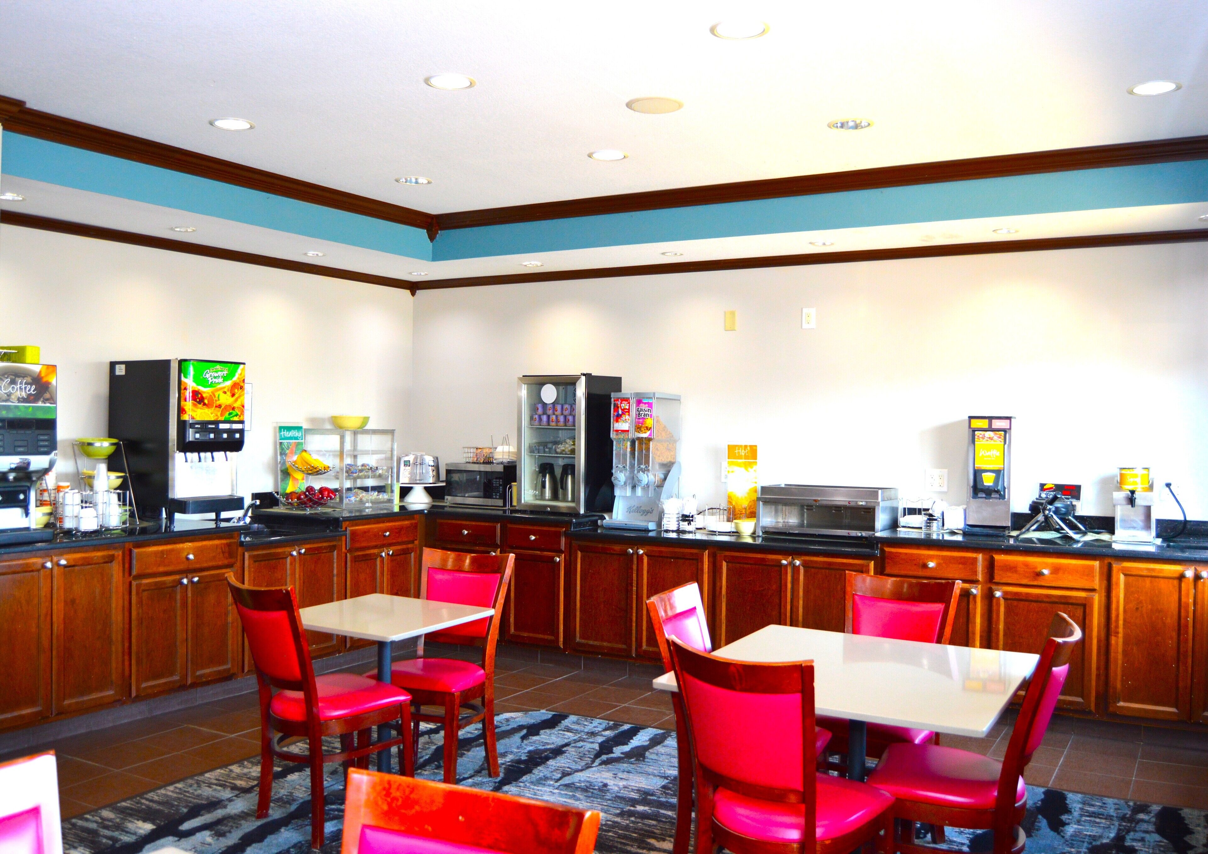 A hotel breakfast area with wooden cabinets, a coffee machine, a microwave, and a refrigerator. There are red chairs and a table with a rug.