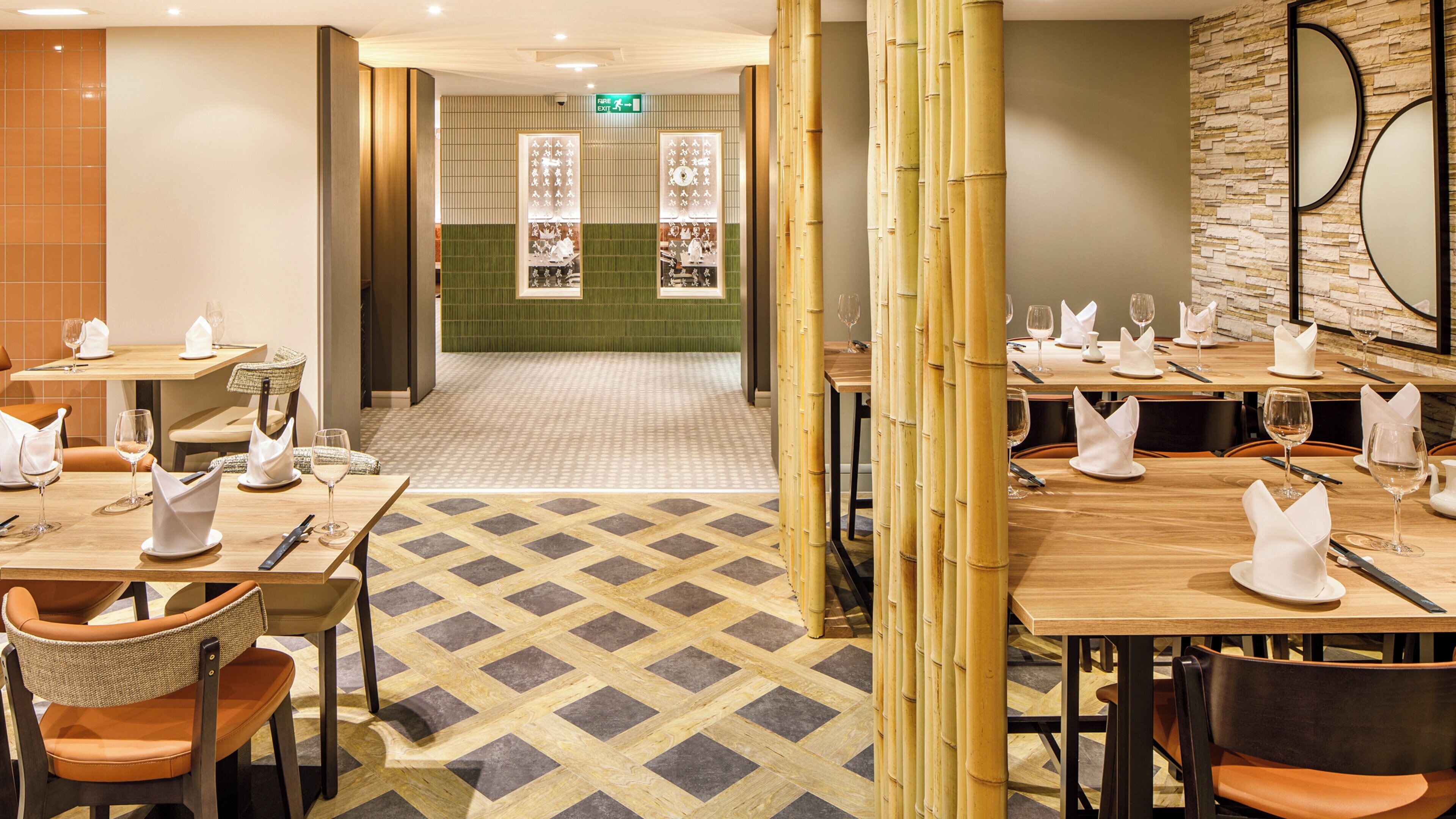 A restaurant with multiple tables set for dining, a green tiled wall, and a bamboo screen.