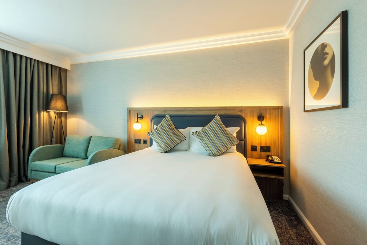 Une chambre d’hôtel avec un grand lit, une table de chevet avec une lampe, une chaise et un tableau encadré au mur.