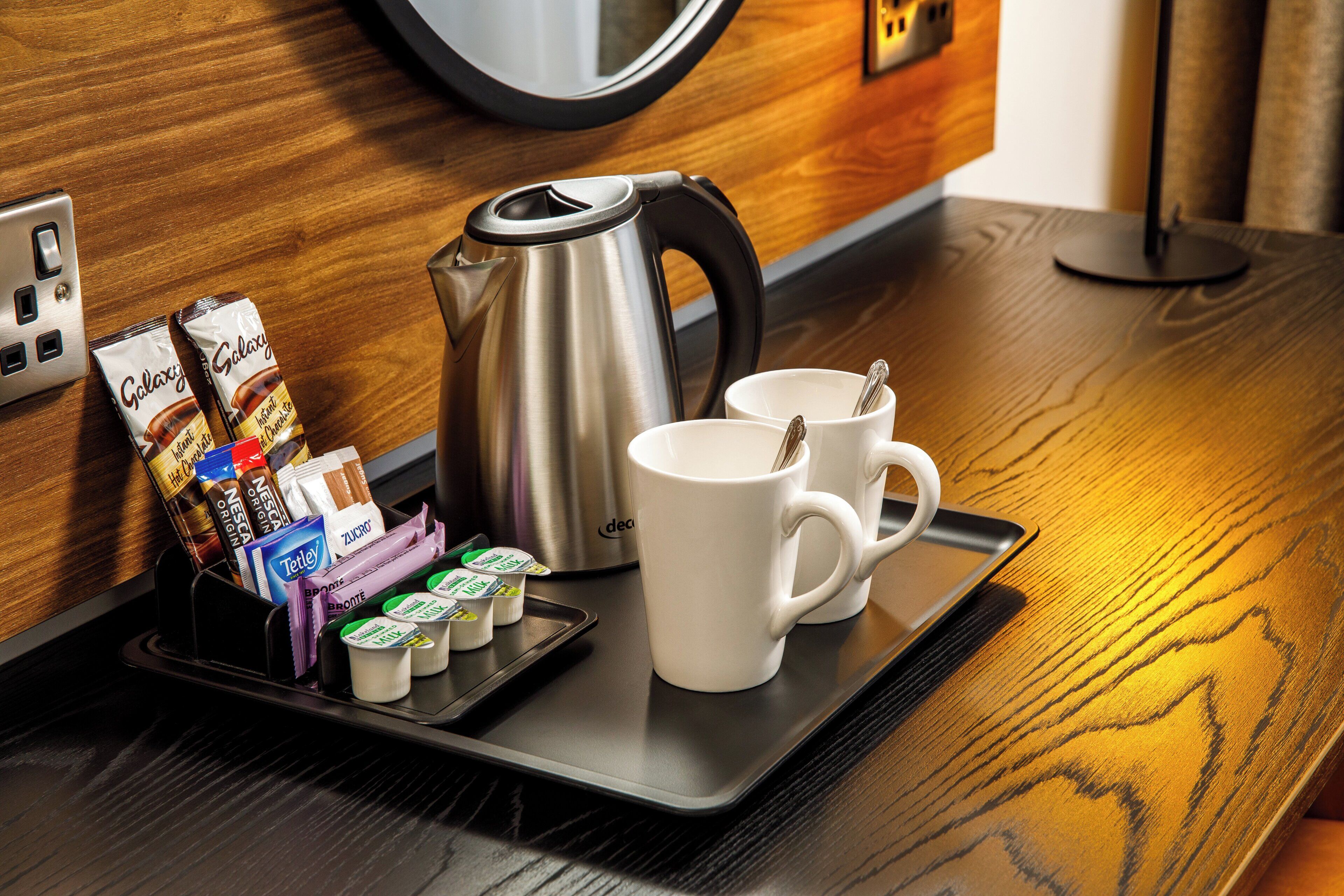 A breakfast tray with a kettle, two white cups, and assorted packaged food items on a wooden table.