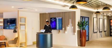 Een hotel receptie met een digitaal scherm, een receptiebalie, zitplaatsen en een vloer met een patroon.