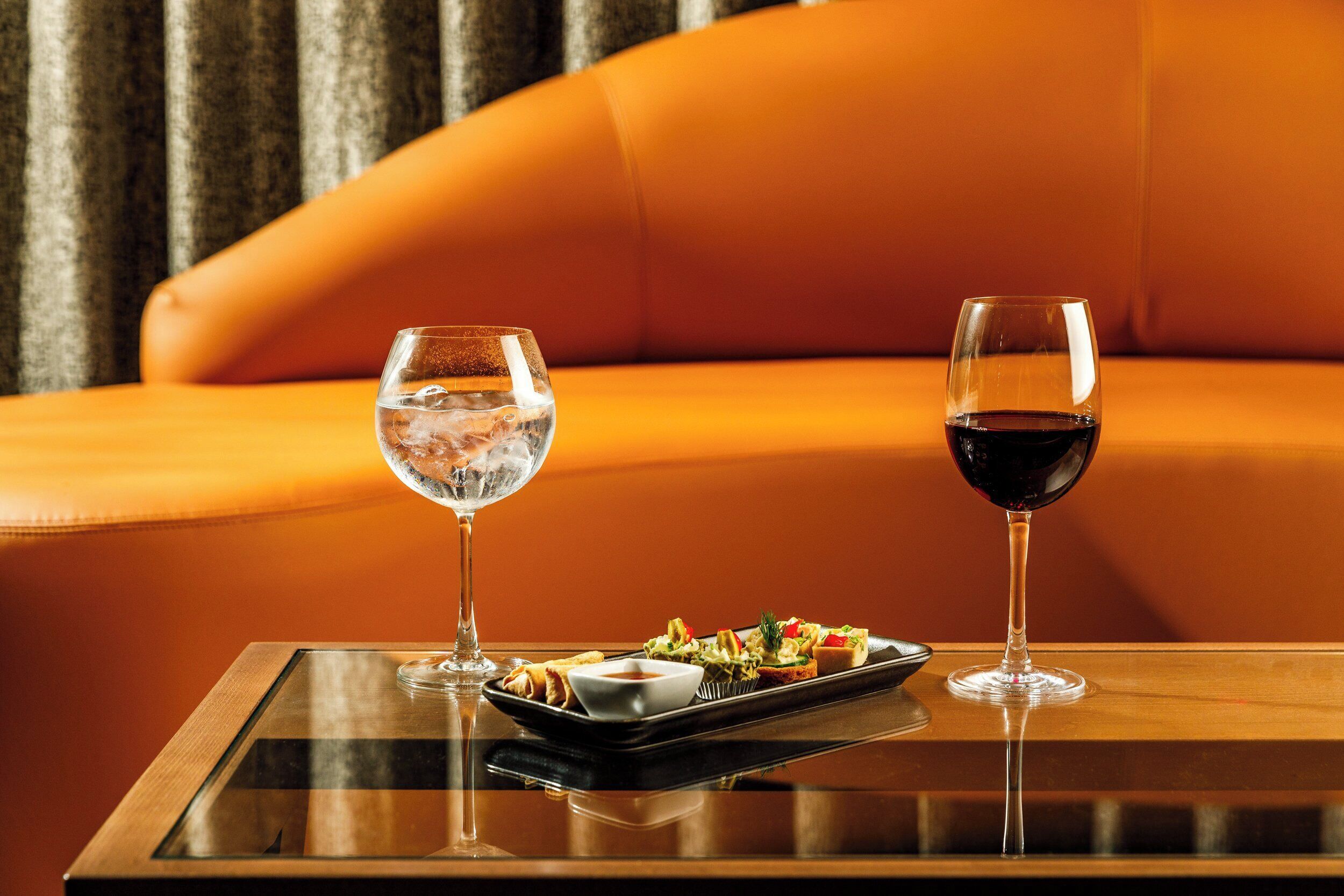 Une table en verre sur laquelle sont disposés un plateau de hors-d'œuvre et deux verres de vin, le tout placé devant un canapé en cuir.