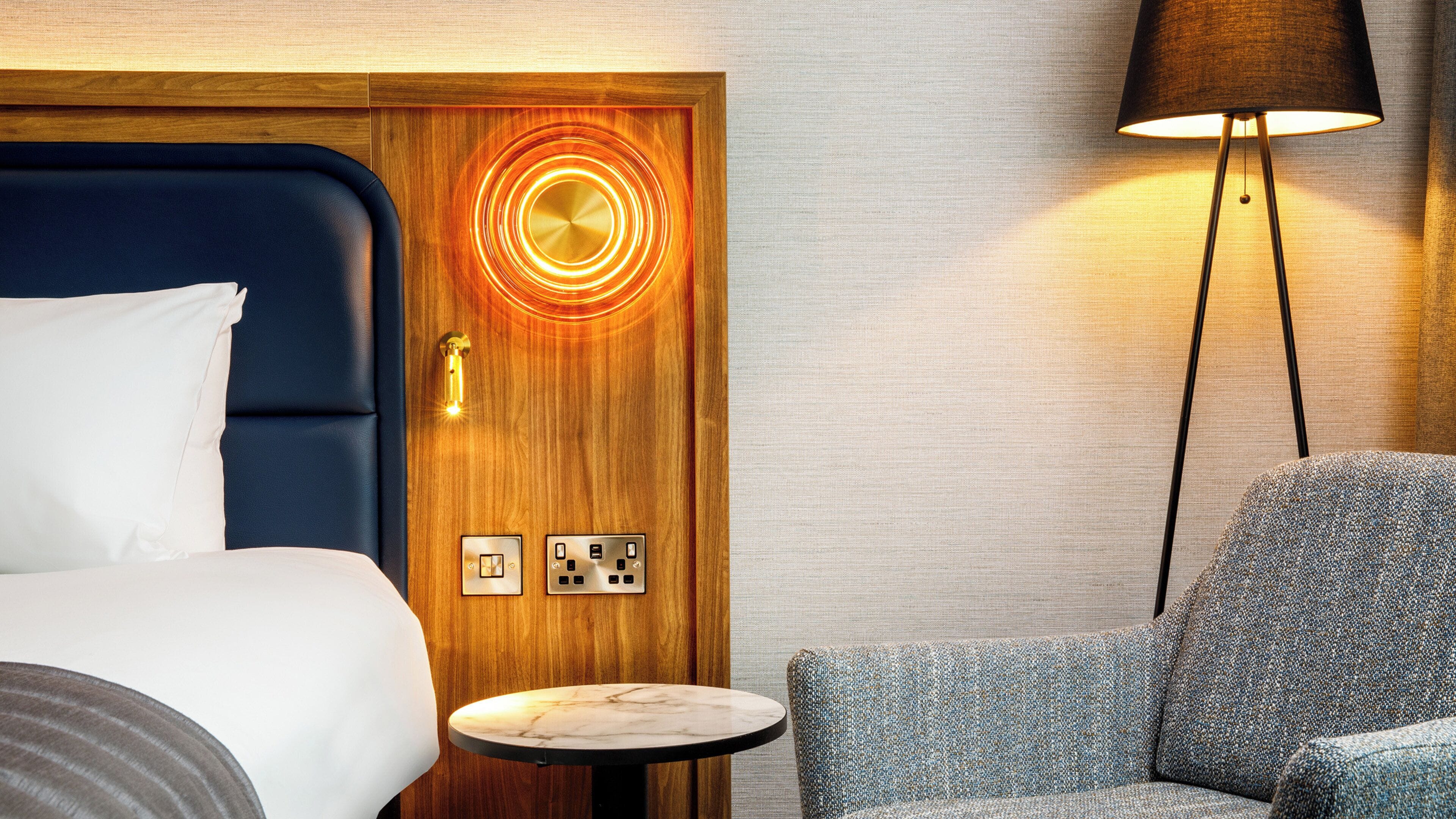 A hotel room with a bed, a small round table, a chair, a lamp, and a door with a circular light design.