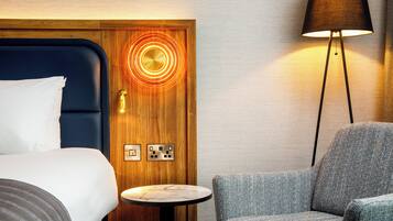 A hotel room with a bed, a small round table, a chair, a lamp, and a door with a circular light design.
