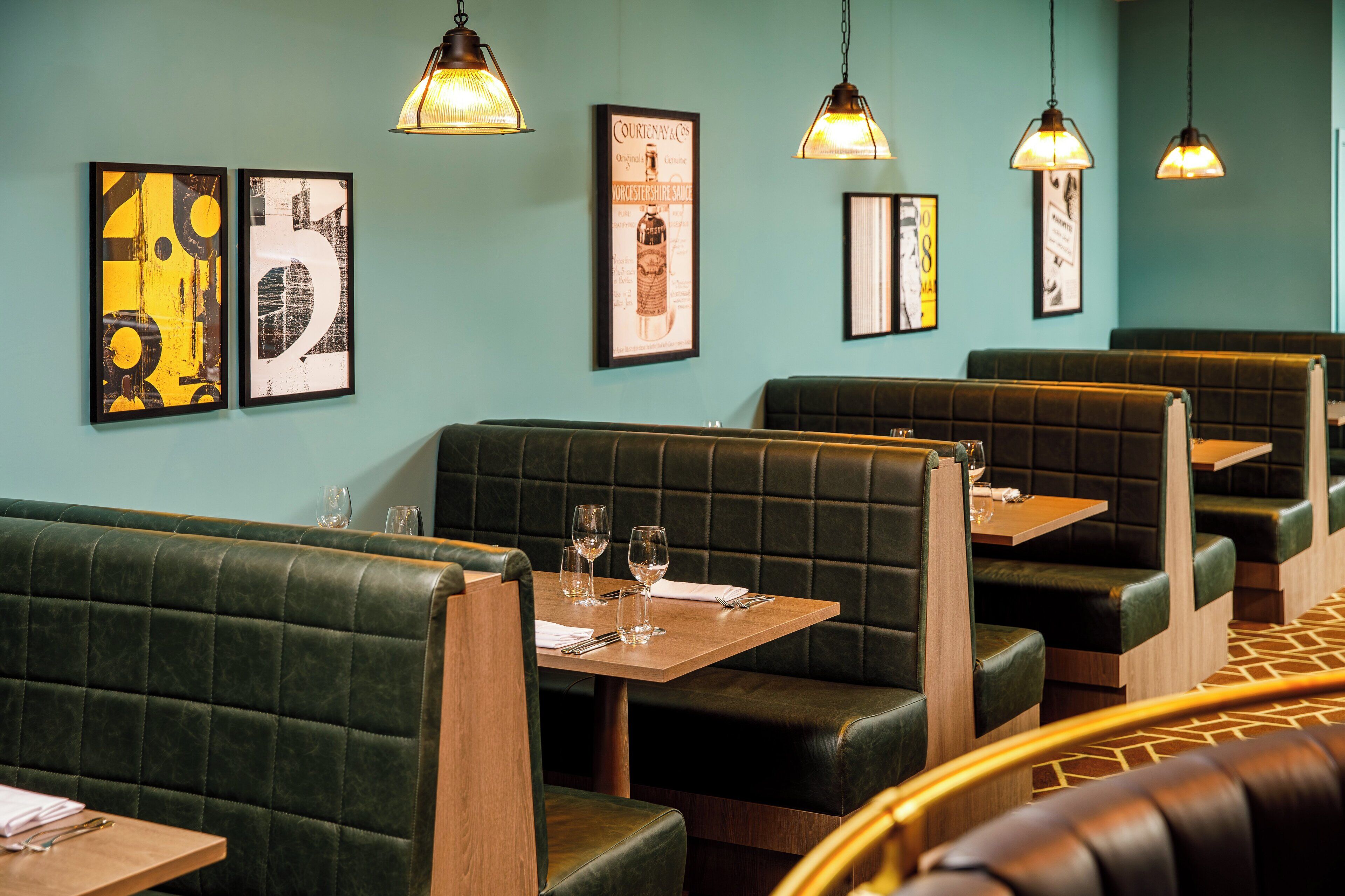 En restaurang med gröna, klädda sittplatser, träbord och hängande taklampor.