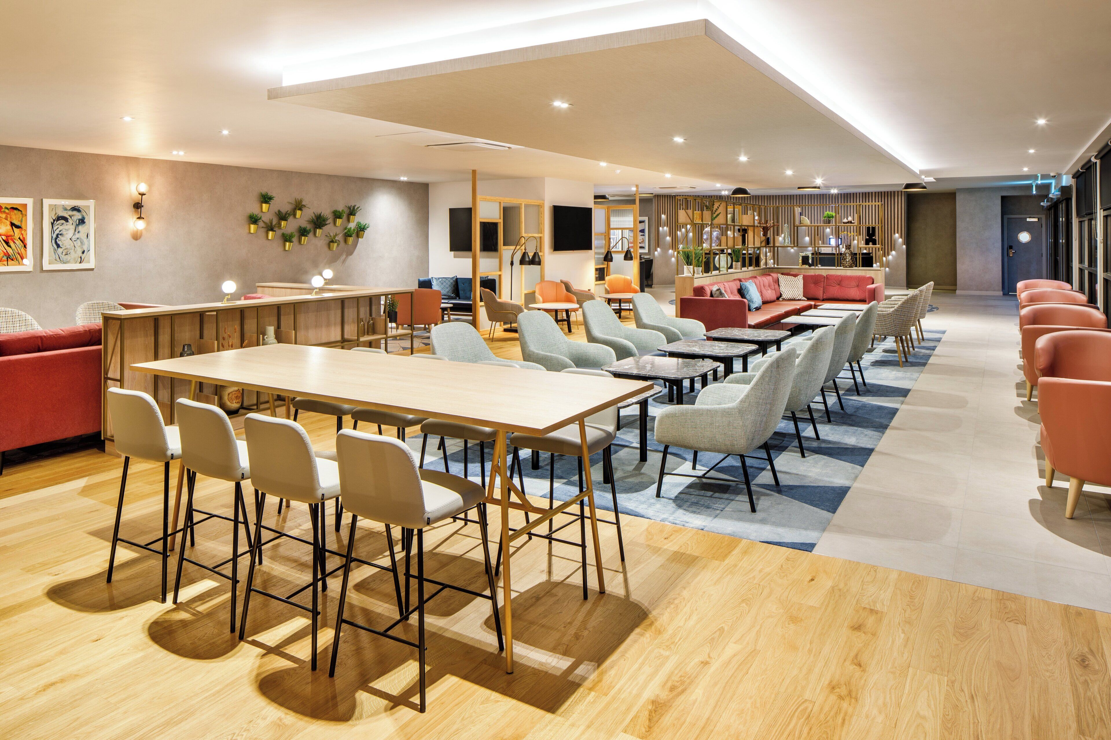 Un moderno vestíbulo de hotel con una larga recepción, varias zonas de estar con sofás y sillones, y un amplio espacio abierto con suelo de madera.