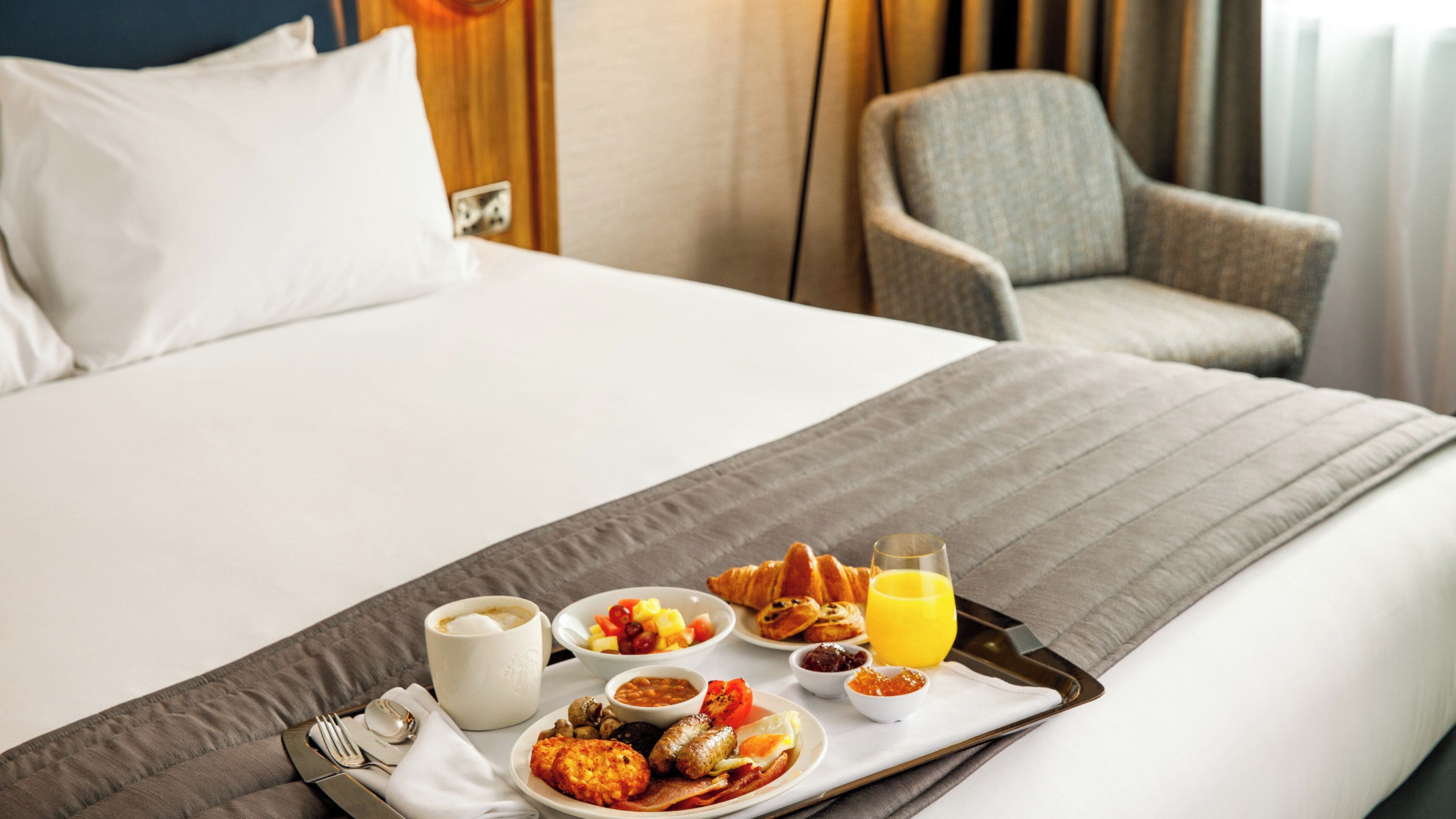 A neatly made breakfast tray on a bed with a variety of food items, including eggs, sausages, and toast, along with a glass of orange juice.