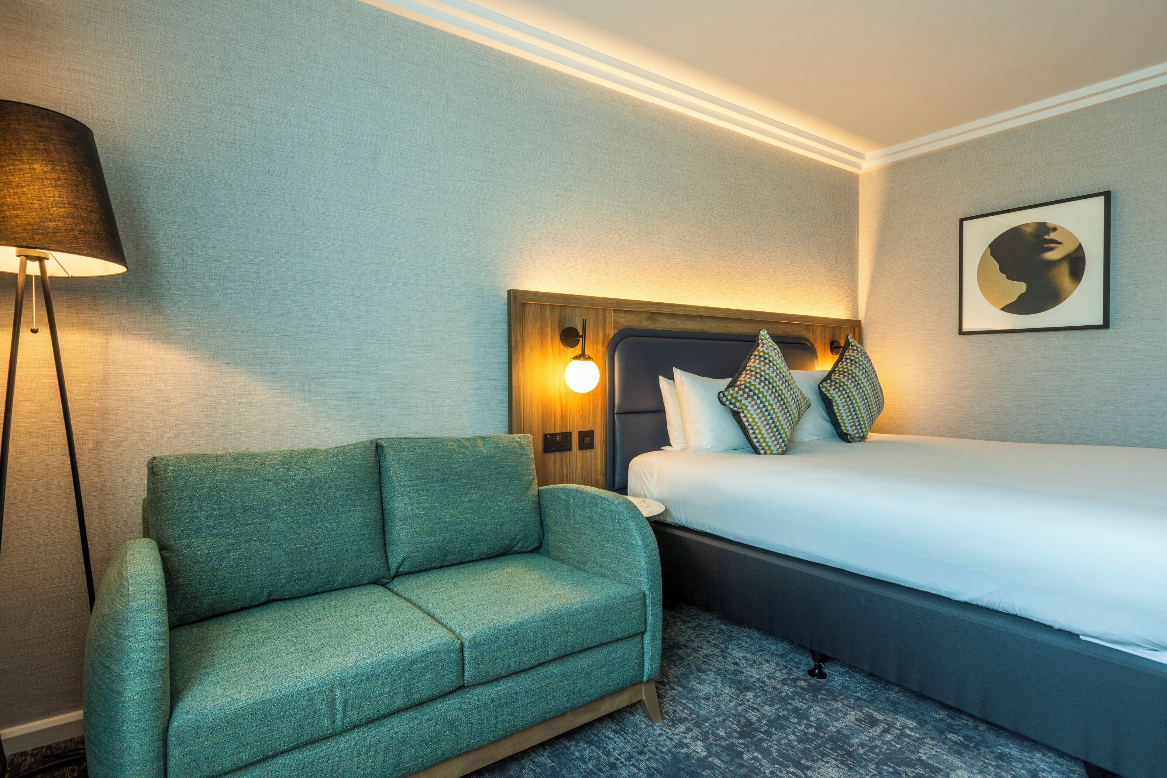 A hotel room with a bed, a green sofa, a floor lamp, and a wall-mounted light fixture.