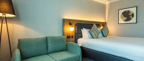 A hotel room with a bed, a green sofa, a floor lamp, and a wall-mounted light fixture.