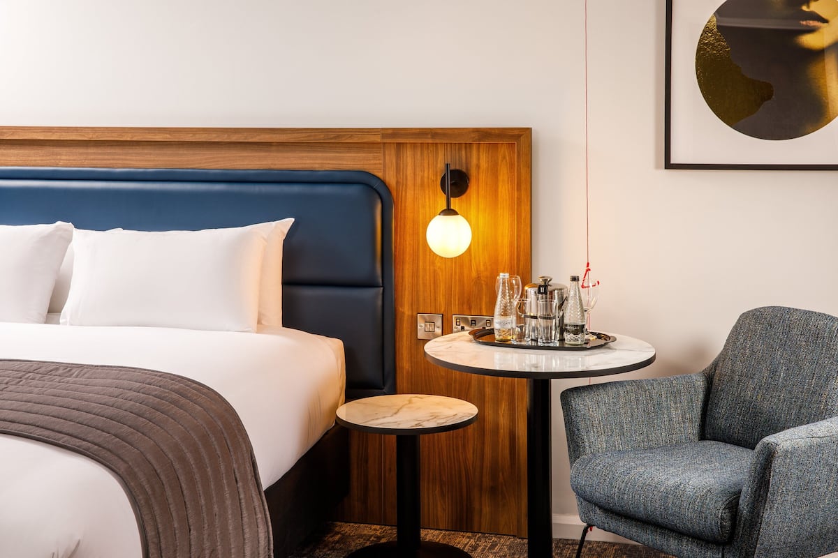 Une chambre d’hôtel avec un lit, une table ronde avec une lampe et un fauteuil bleu.