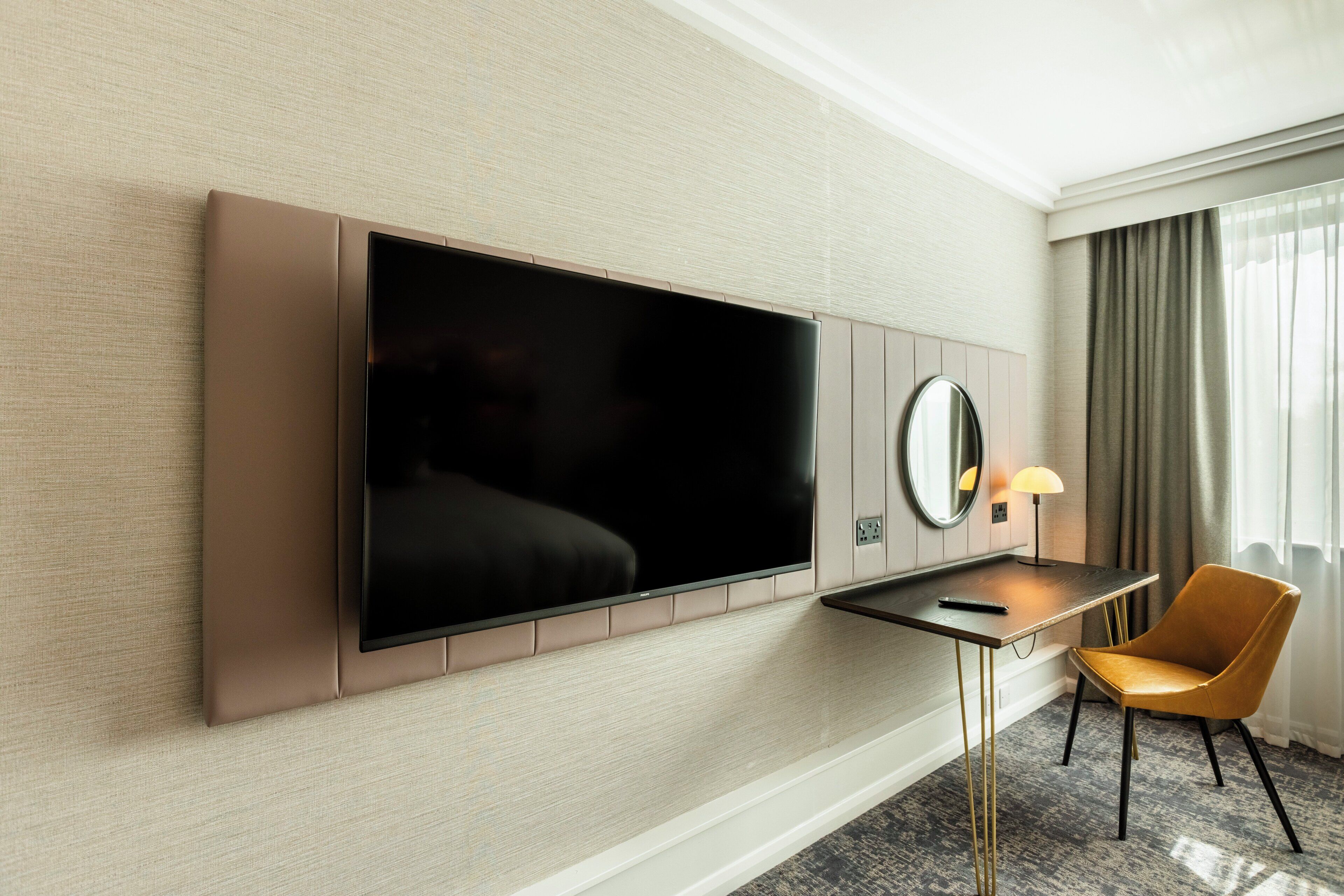 A hotel room with a flat-screen TV mounted on the wall, a small desk with a lamp, a chair, and a large mirror.