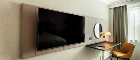A hotel room with a flat-screen TV mounted on the wall, a small desk with a lamp, a chair, and a large mirror.