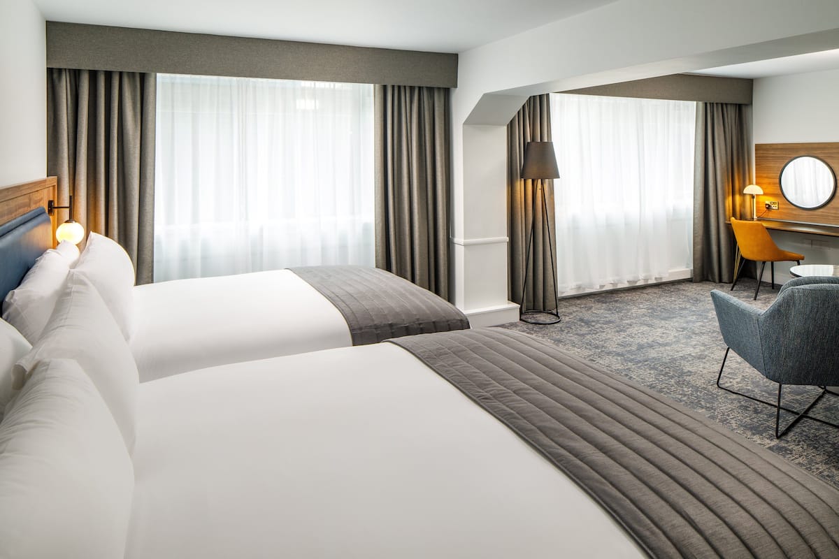 Une chambre d’hôtel avec deux lits, un bureau avec une chaise, une lampe et une grande fenêtre avec des rideaux.