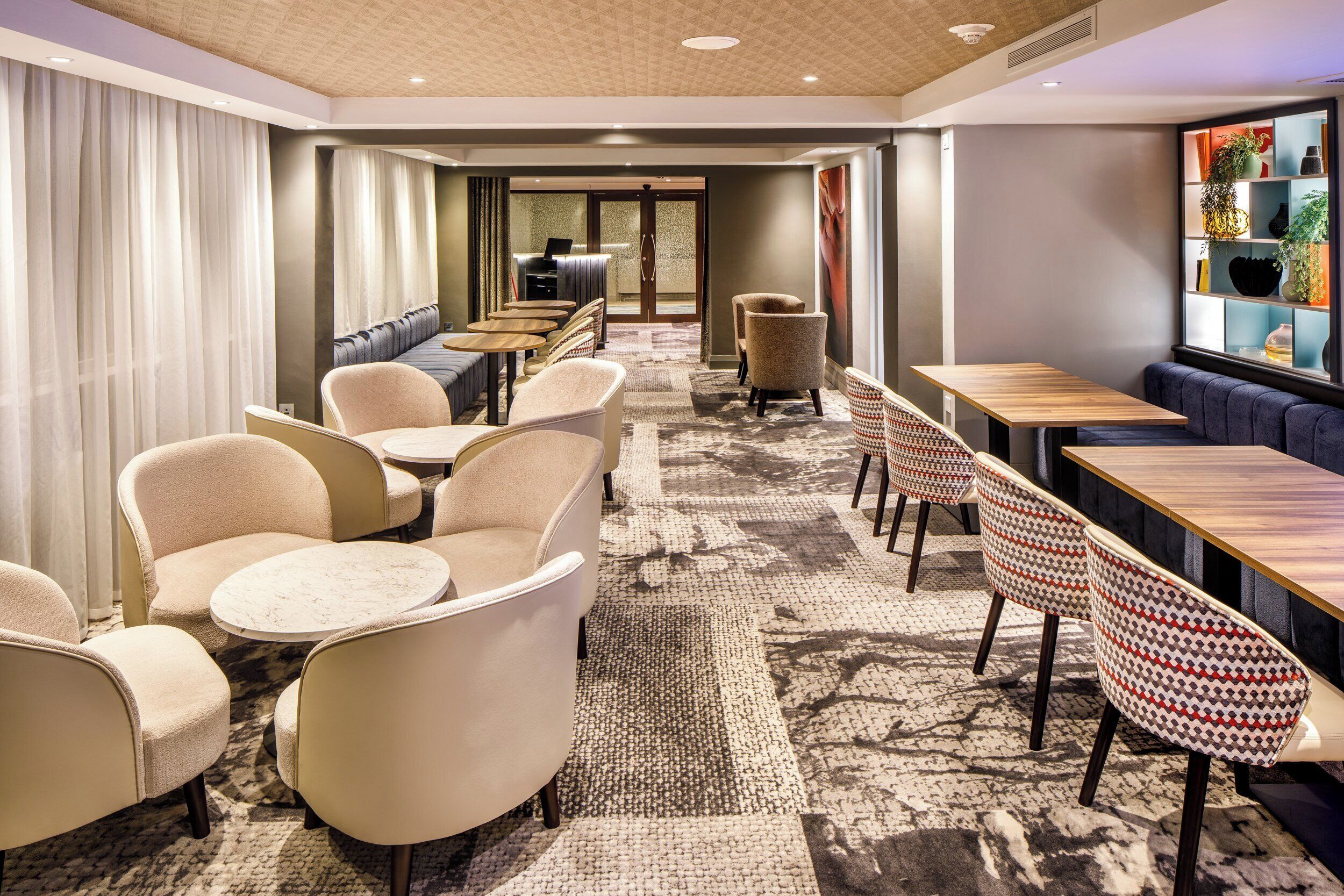 Un hall d’hôtel doté d’un long comptoir d’accueil, de plusieurs espaces aménagés avec des tables et des chaises, d’un tapis à motifs et d’une grande fenêtre offrant une vue panoramique.