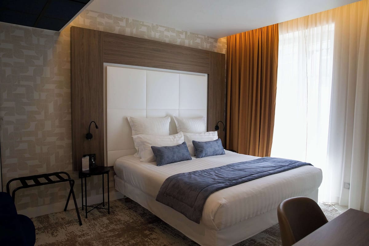 A modern hotel room with a large bed, a bedside table, a chair, and a view of the outdoors through the window.