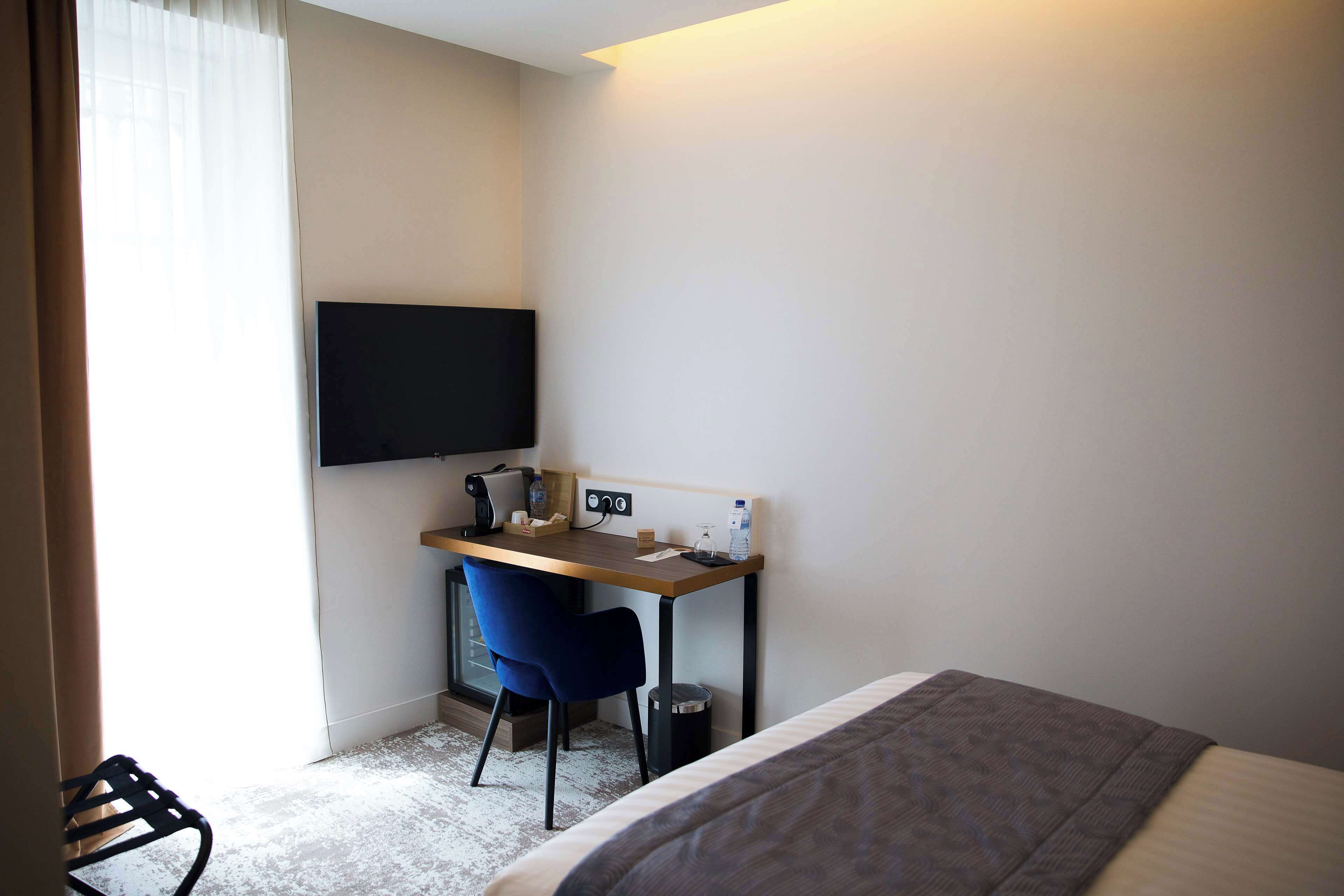 A hotel room with a bed, a desk with a chair, a flat-screen TV, a coffee machine, and a trash bin.